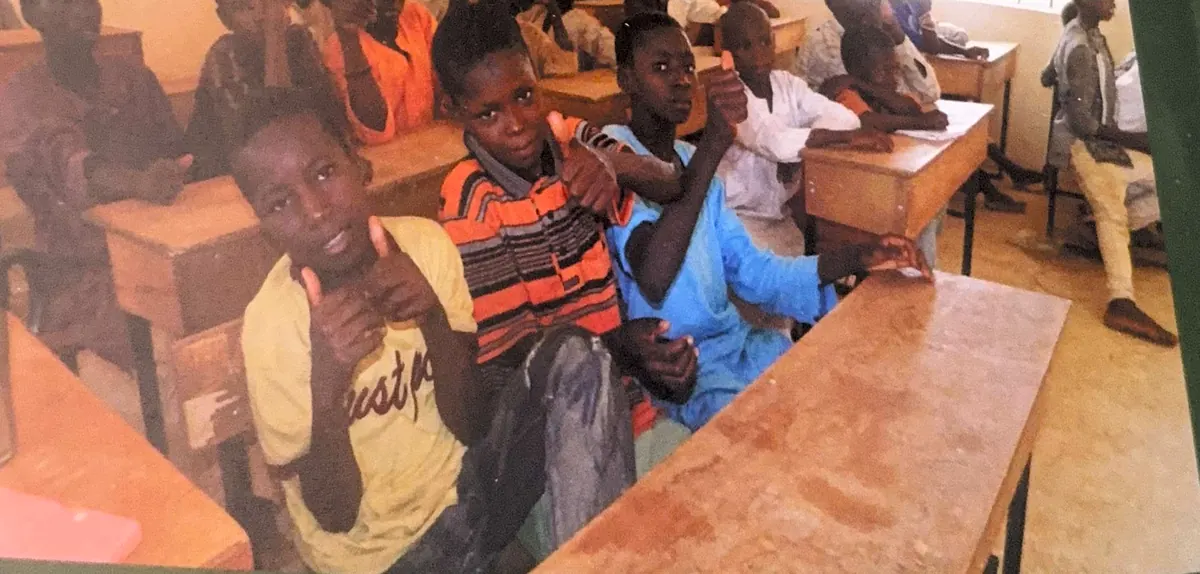 Pupils sitting at wooden desks in a classroom smile and give a thumbs-up to the camera.