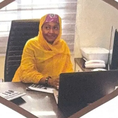 Maryam Umar Aliyu, wearing a yellow hijab, smiles from behind her office desk.