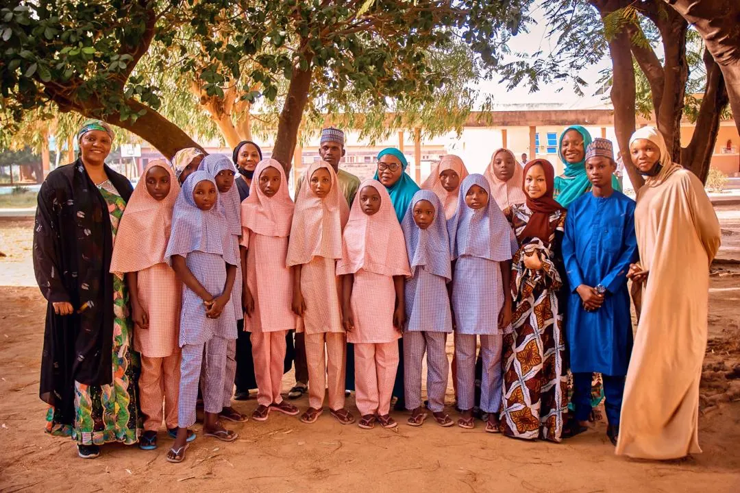A group of schoolgirls in pink and blue gingham uniforms and matching hijabs stand in a row beneath shady trees, with women and young men in traditional dress on either side.