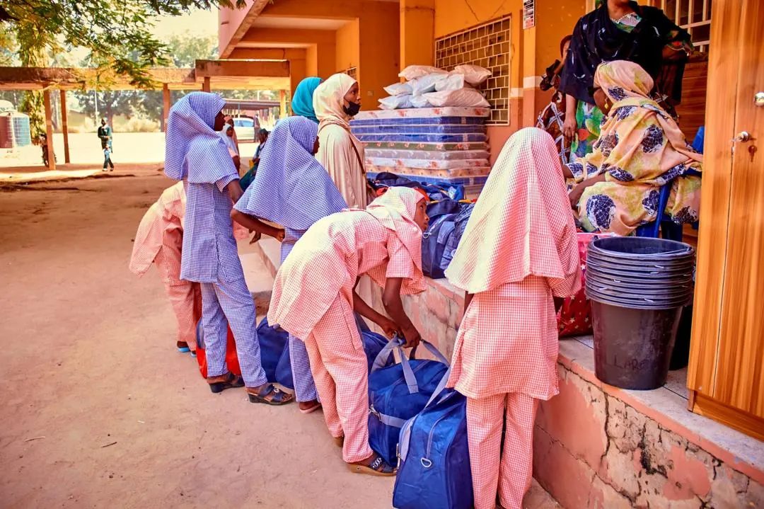 Schoolgirls in gingham uniforms and hijabs bend over blue travel bags outside a school building, beside a stack of mattresses and a pile of buckets.